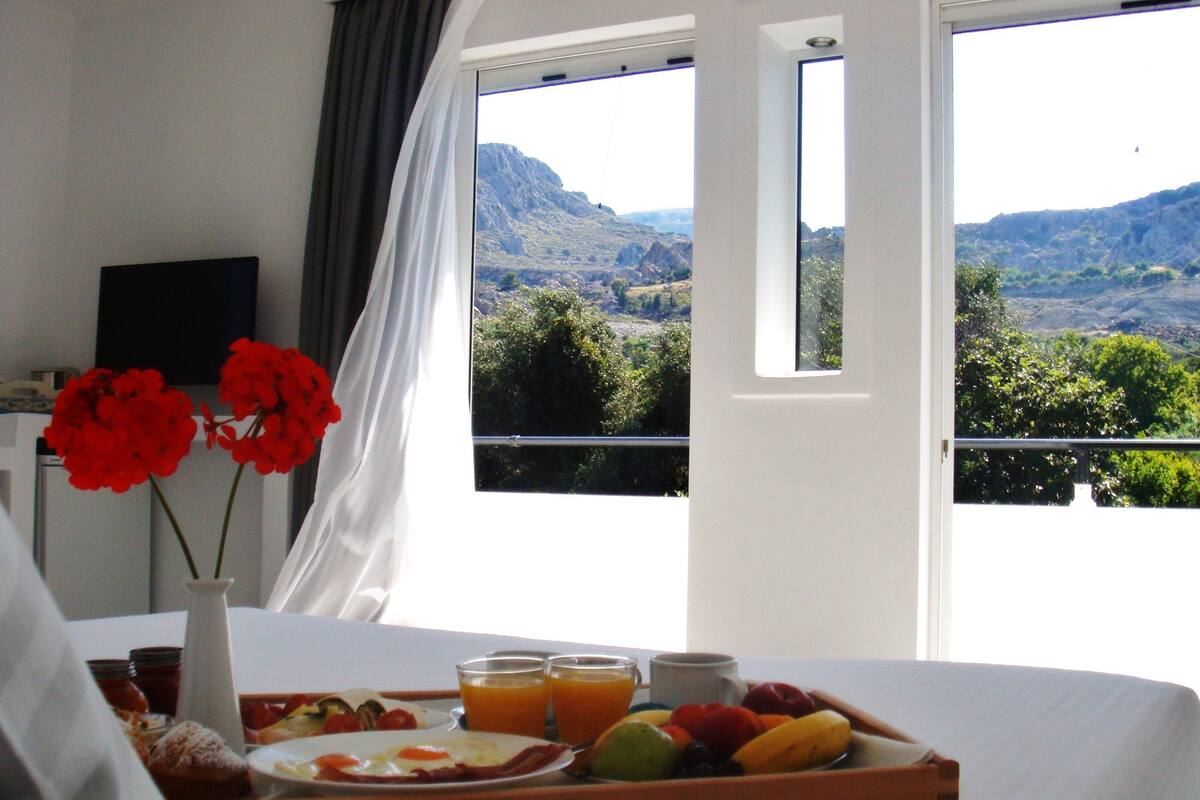 Bandeja de café da manhã com frutas, suco e um prato de ovos em uma cama, com vista para uma paisagem montanhosa através de grandes janelas.