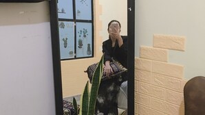 A person taking a selfie in a room with a patterned sofa, a large plant, and a window with a geometric wall design. - Hani House (Pleiku)