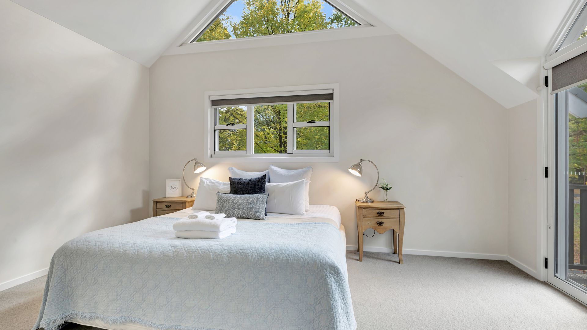 A bedroom with a skylight, a bed with a white duvet and pillows, two bedside tables with lamps, and a large window with a view of trees.