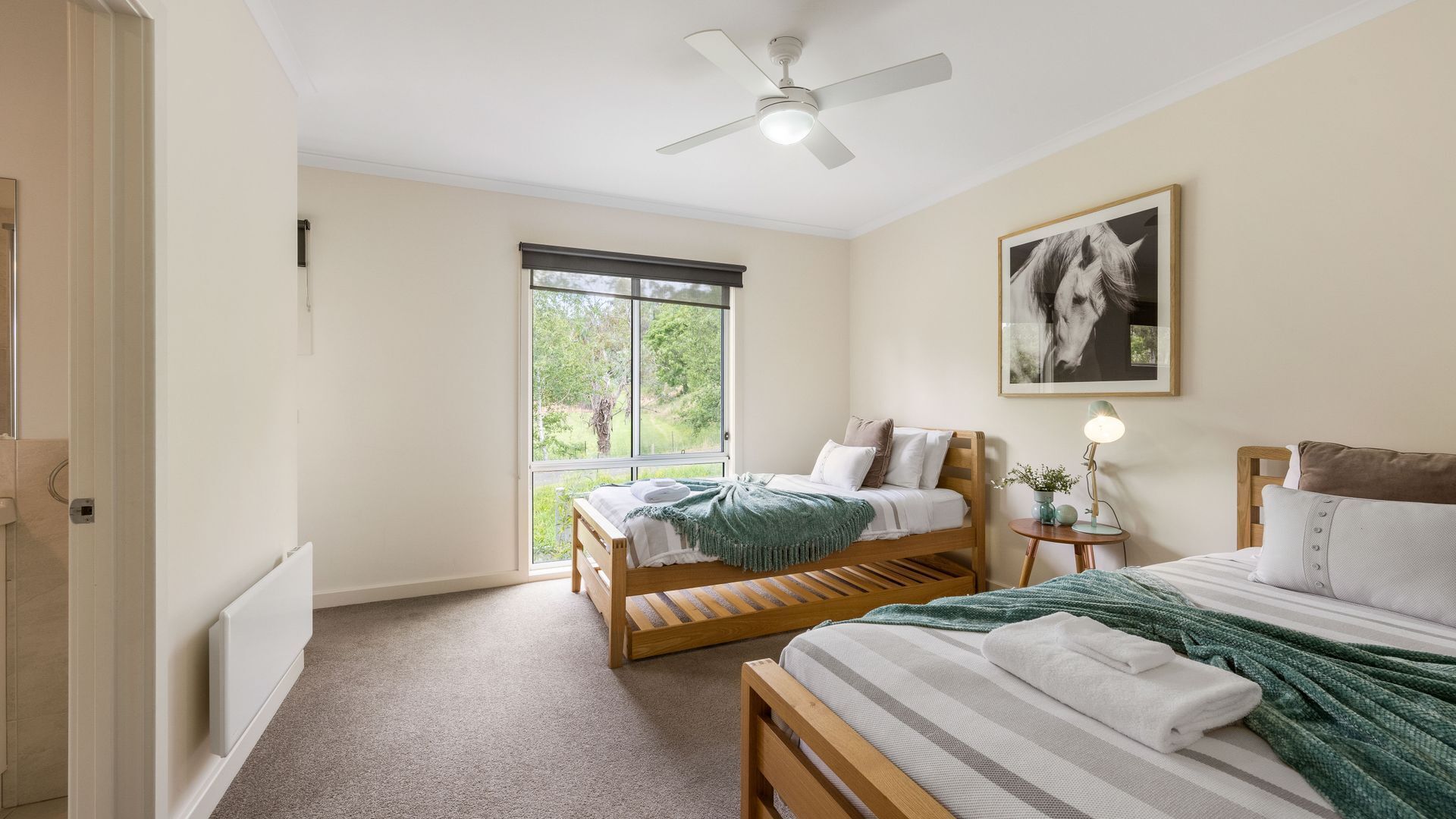 A bedroom with two wooden beds, a ceiling fan, a large window with a view of greenery, and a framed picture on the wall.