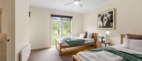 A bedroom with two wooden beds, a ceiling fan, a large window with a view of greenery, and a framed picture on the wall.