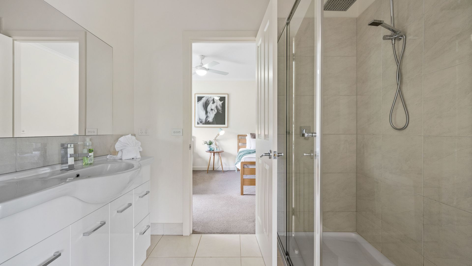 A modern bathroom with a glass shower enclosure, a white vanity with a mirror, and a door leading to a bedroom.