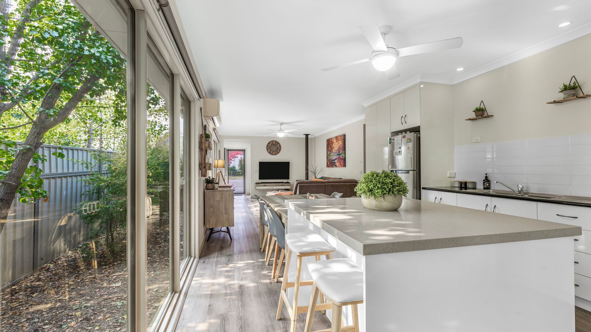 A modern kitchen with a large island, bar stools, a dining area, and a view of a tree outside.