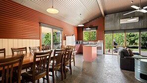 A modern kitchen with a central island, wooden dining table, and chairs. There is a view of greenery outside through large windows. - Quins Gap Escapes (Bright)