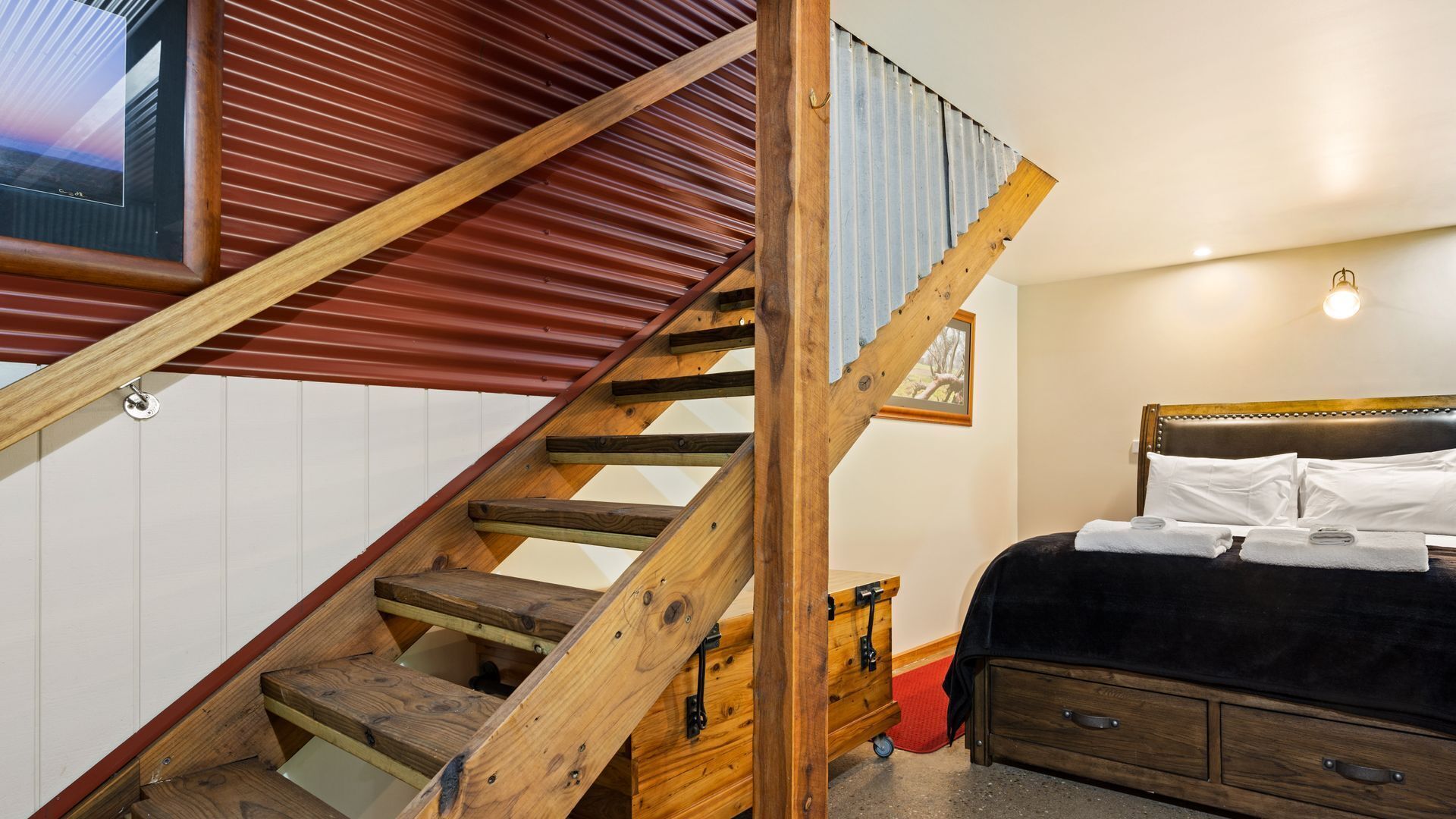 A bedroom with a wooden staircase, a bed with white linens, and a framed picture on the wall.