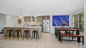A modern kitchen with a central island, bar stools, a dining table with chairs, and a large window with blinds. - Always Bright - A Bright Escape (BRIGHT)
