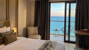 Habitación de hotel con cama, una silla, una mesita y vista al océano a través de amplias puertas correderas.