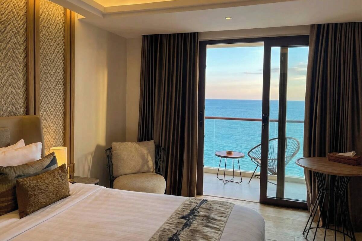 Habitación de hotel con cama, una silla, una mesita y vista al océano a través de amplias puertas correderas.