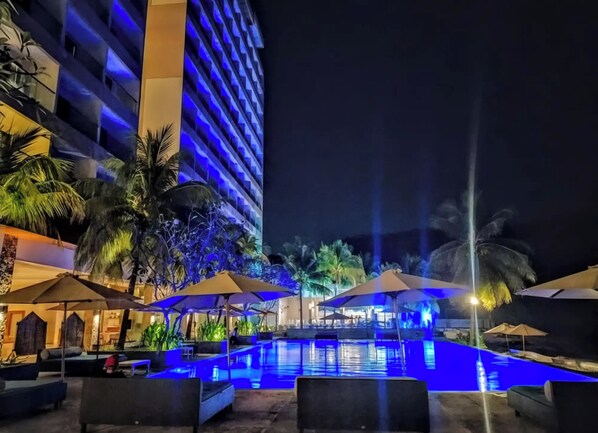 Vue nocturne d’une piscine d’hôtel, avec un grand bâtiment en arrière-plan, des palmiers et des chaises longues.