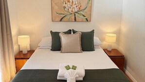 Una cama bien hecha con una manta verde, dos almohadas y una toalla doblada. Dos lámparas de noche sobre mesitas de madera. Un cuadro cuelga sobre la cama.