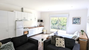 Un apartamento moderno con cocina, dos sofás negros, una mesita y un amplio ventanal con vistas al verdor.