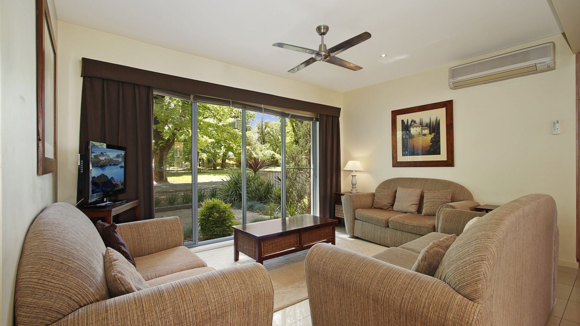 A living room with a large sliding glass door, a ceiling fan, a television, and a painting on the wall.