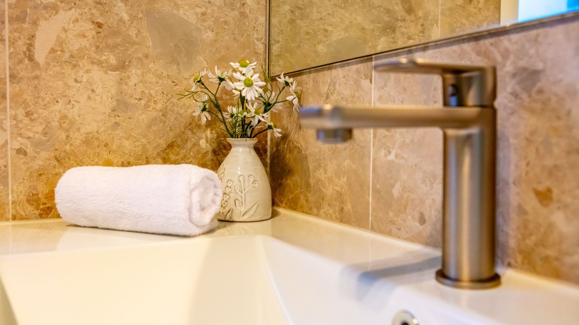 A bathroom with a marble wall, a white sink, a white towel, and a vase with flowers.
