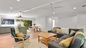 A modern living room with a sofa, armchair, and coffee table. There is a dining area with a table and chairs. The room has a ceiling fan and recessed lighting.