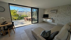 Un salon moderne doté d’une porte-fenêtre coulissante donnant sur une terrasse extérieure, d’un téléviseur à écran plat fixé au mur et d’un canapé beige agrémenté de coussins.