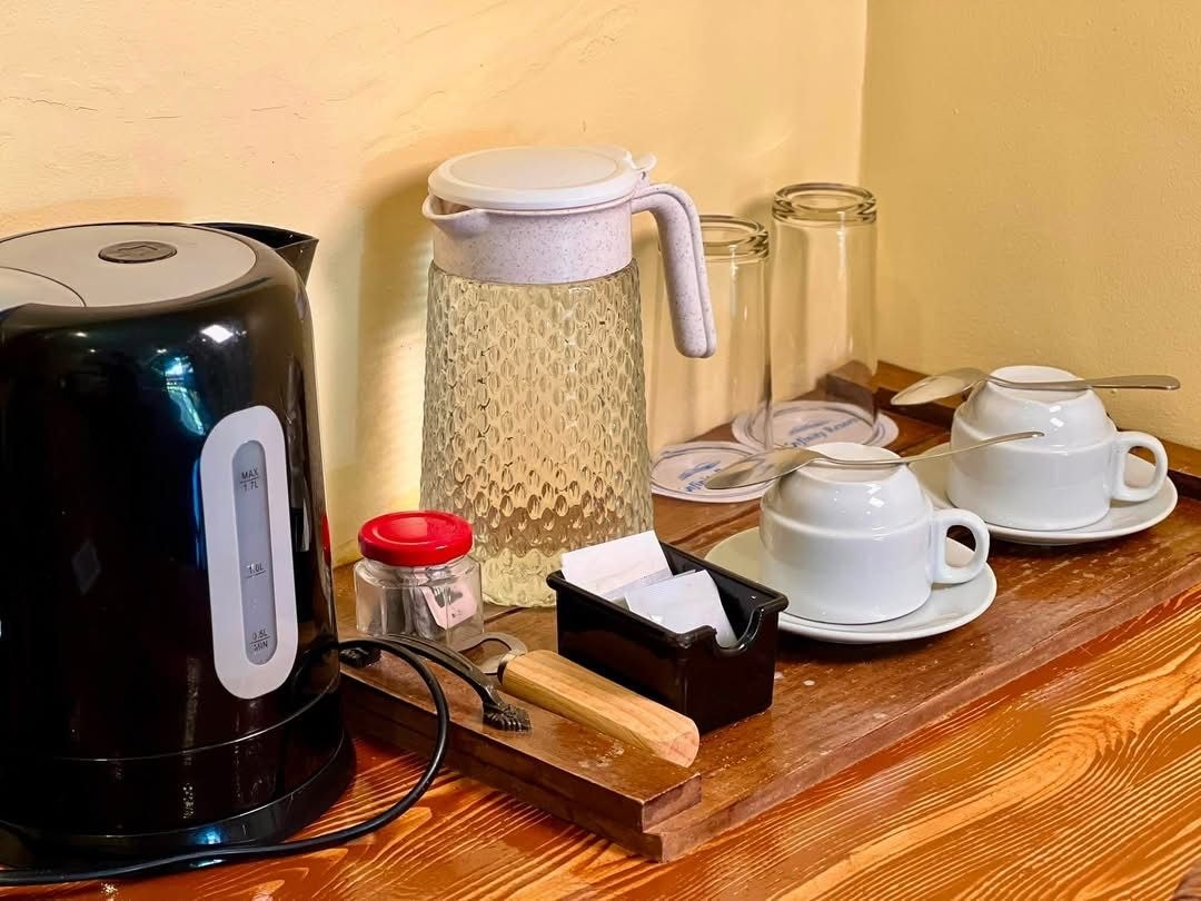 A wooden table with a black electric kettle, a textured glass carafe, white teacups, a small container with a red lid, and a tissue box.
