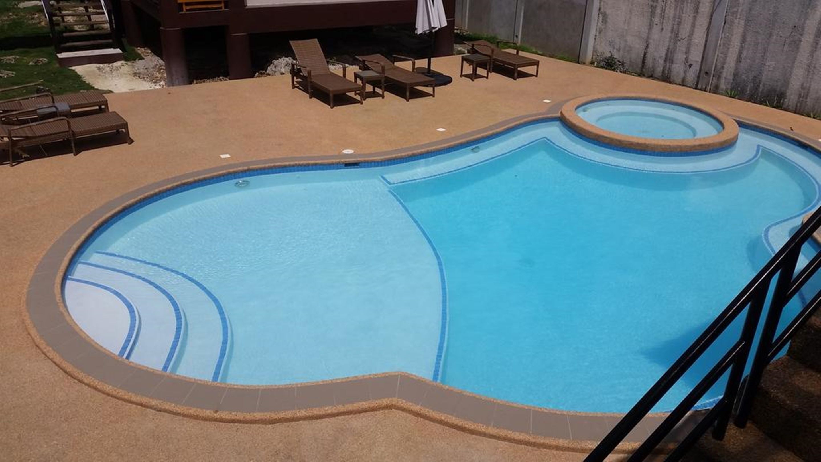 A swimming pool with a curved design, surrounded by a sandy deck and equipped with lounge chairs.
