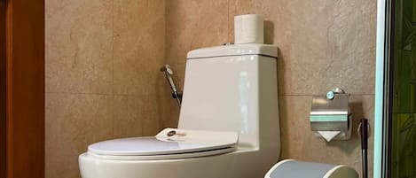 Un baño con inodoro, un cubo de basura y dispensador de toallas de papel en la pared.