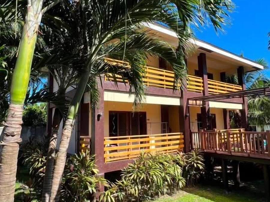 Un edificio de dos pisos con un balcón, rodeado de palmeras y un césped bien cuidado.