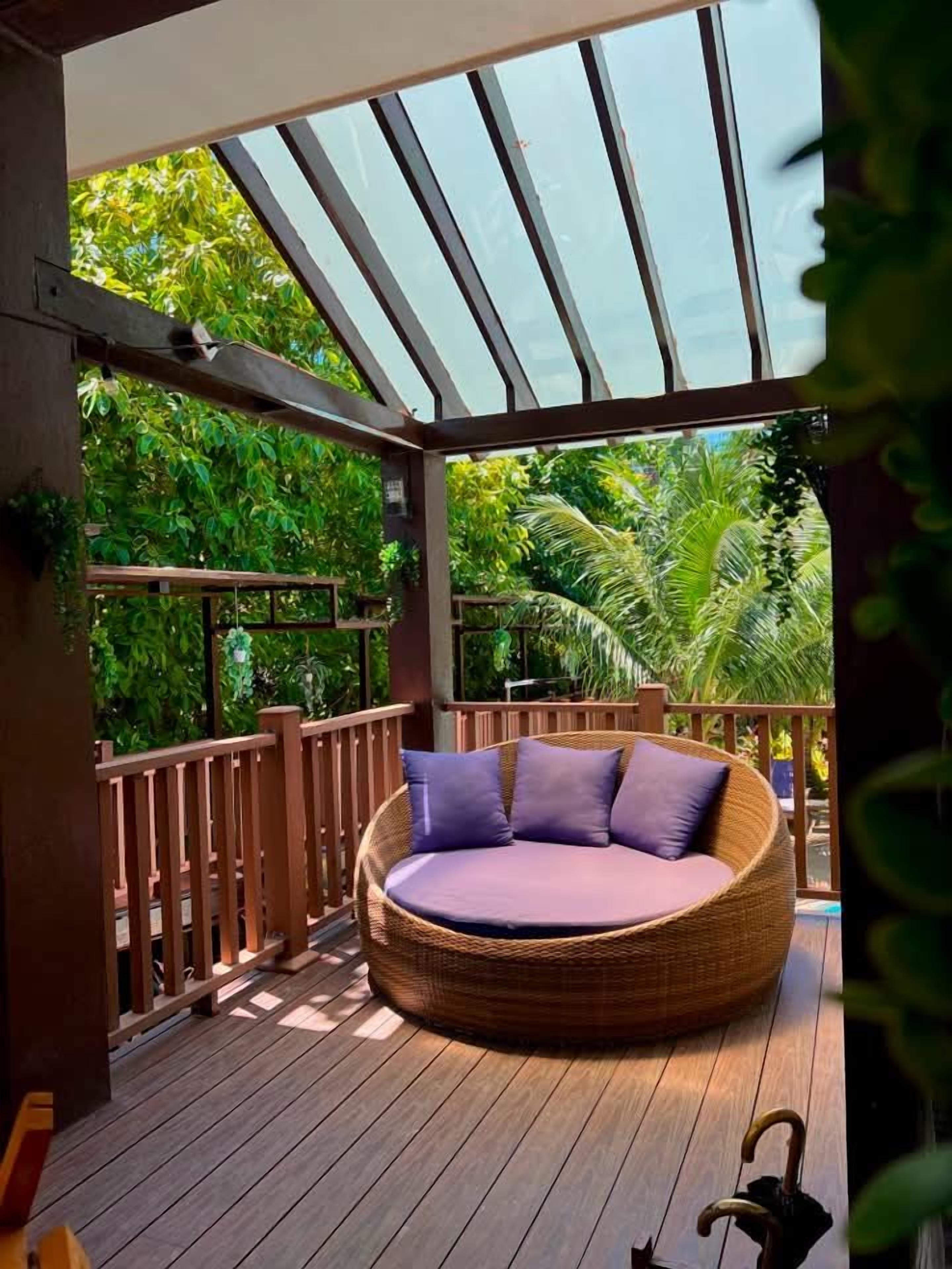 A covered outdoor seating area with a round wicker sofa and cushions, surrounded by wooden railings and lush greenery.