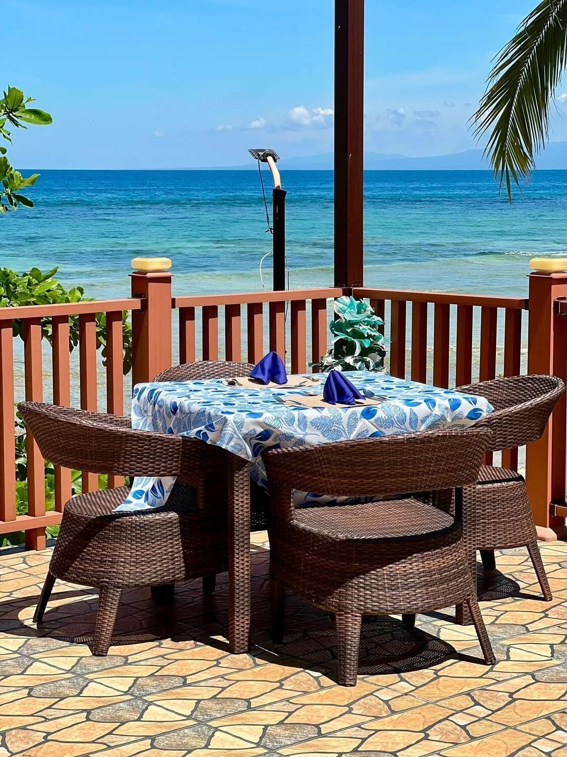 A wicker table set with a blue and white tablecloth, surrounded by wicker chairs, on a patio with a view of the ocean.