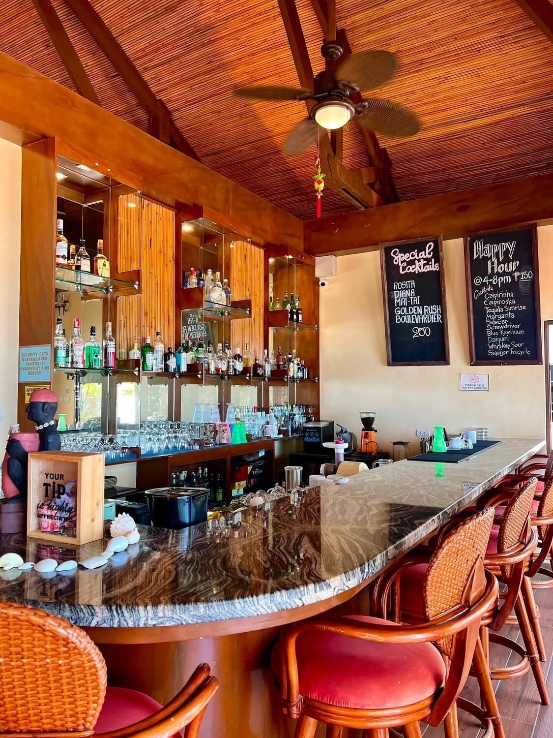 Un área de bar con techo de madera, mostrador central y sillas de mimbre.