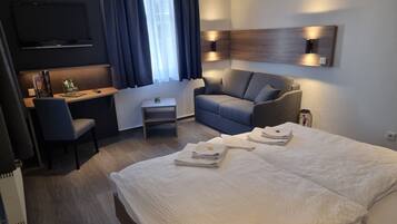 A hotel room with a bed, a desk, a chair, a sofa, and a TV mounted on the wall.