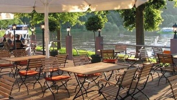 An outdoor patio with wooden tables and chairs, surrounded by a brick floor and greenery.