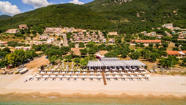 A coastal town with a beach area featuring rows of sun loungers and umbrellas, surrounded by residential buildings and a mountainous backdrop. - Akti Barbati Houses by Konnect (Corfu)