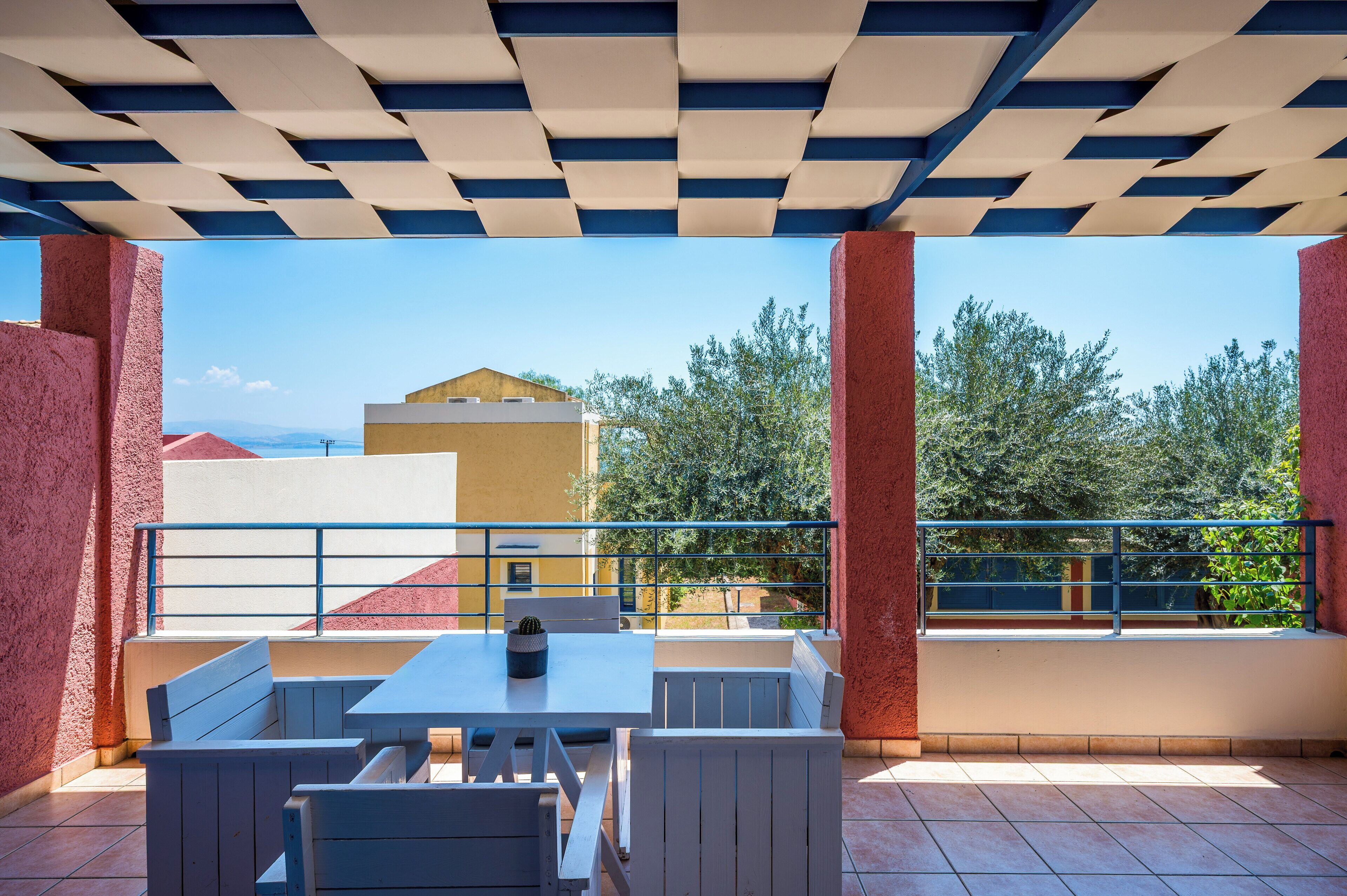 Un espacio de estar al aire libre cubierto con una mesa, bancos, barandilla y vistas a edificios y árboles.