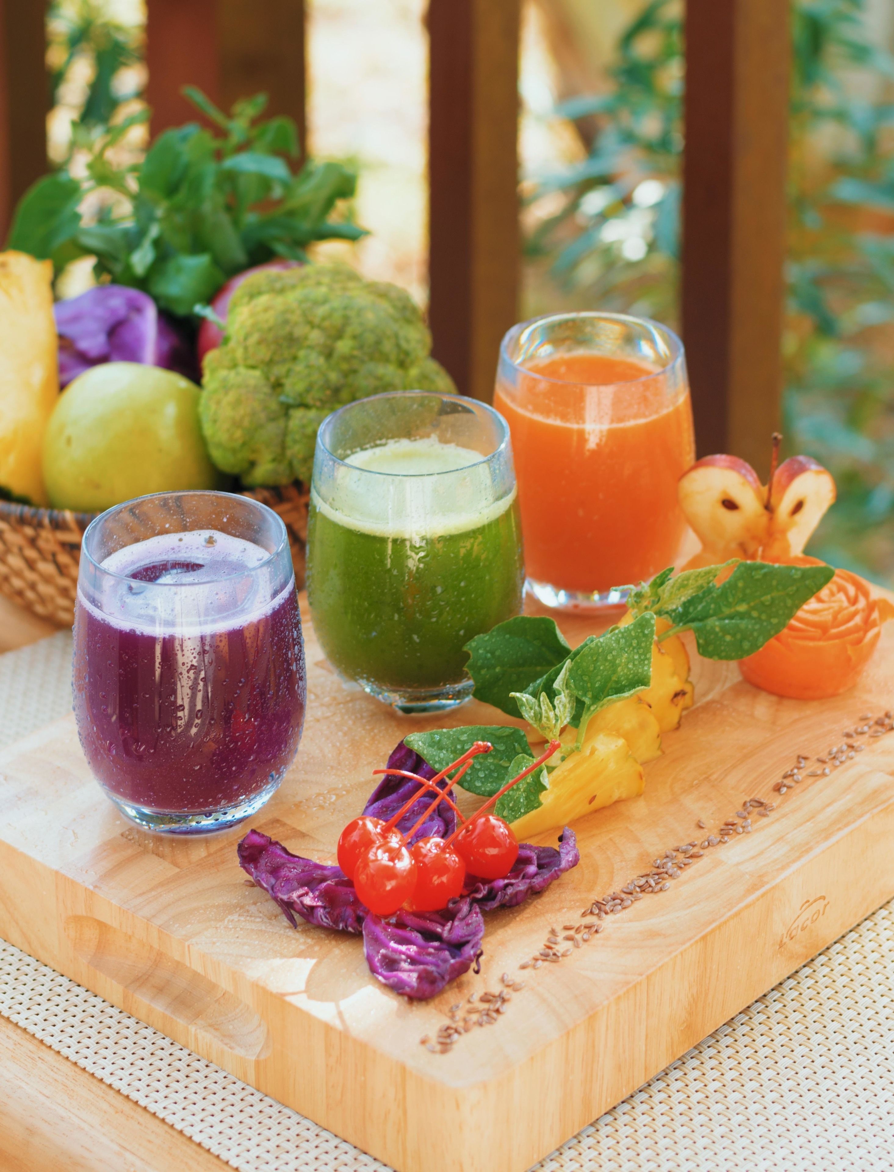 Una mesa de madera con tres vasos de bebidas de colores distintos, una variedad de frutas y un arreglo decorativo de frutas.
