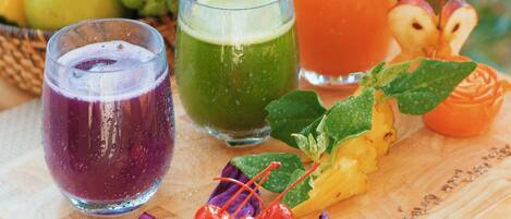 A wooden table with three glasses of different colored beverages, a variety of fruits, and a decorative fruit arrangement.
