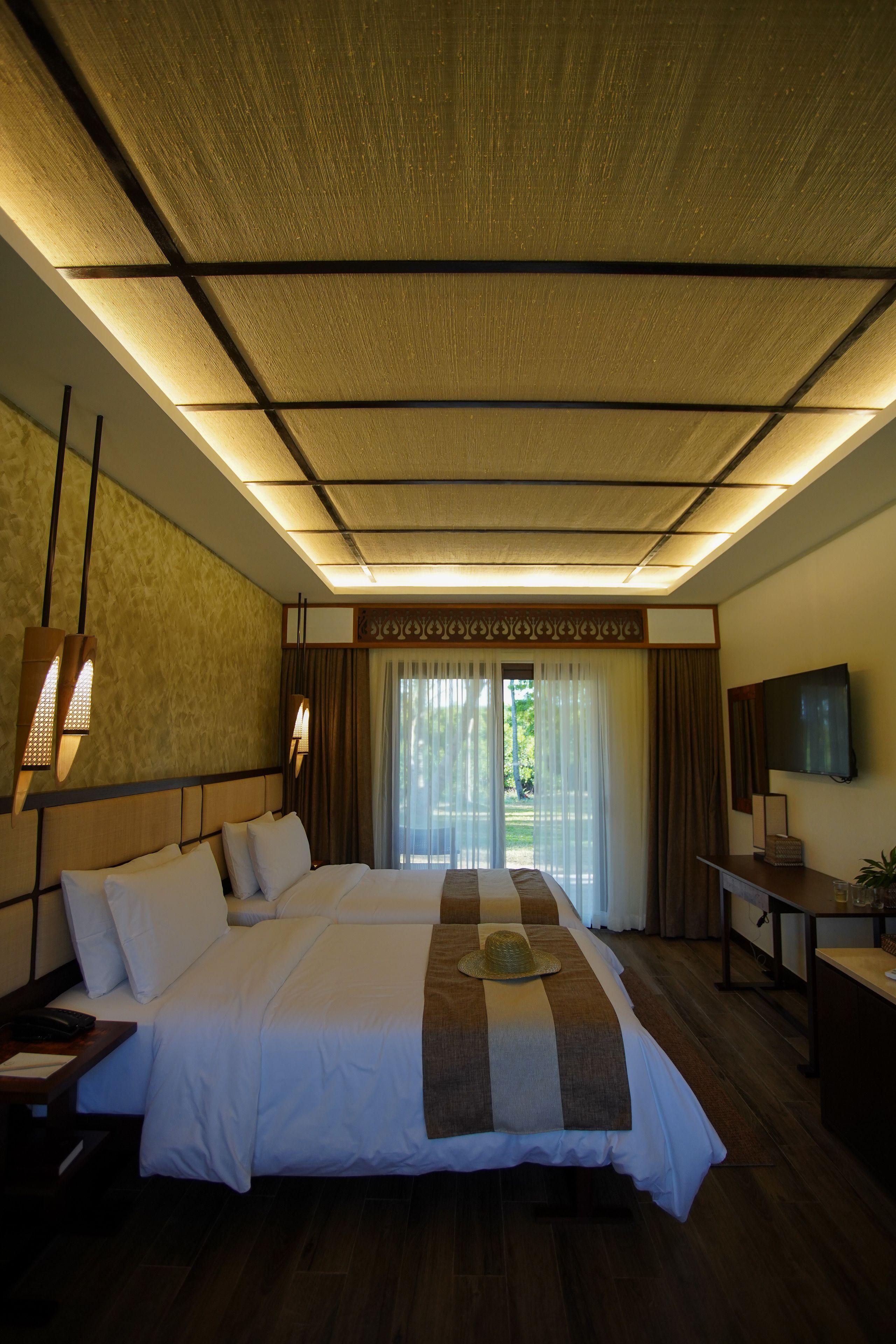 A hotel room with a large bed, a flat-screen TV, a desk with a lamp, and a view of greenery through the window.