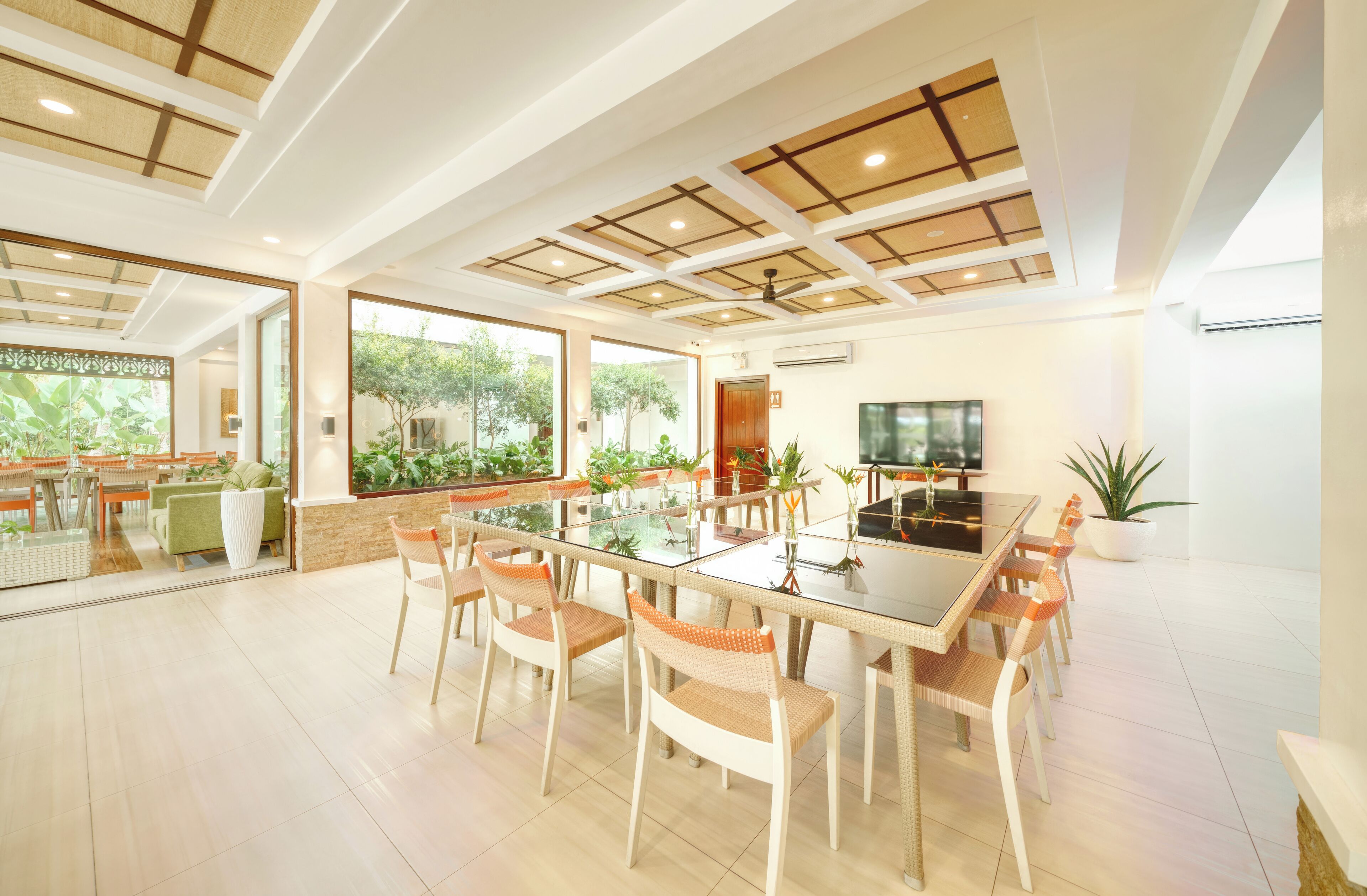 A spacious dining area with a glass table and wicker chairs, large windows offering views of greenery, and a ceiling with a grid pattern.