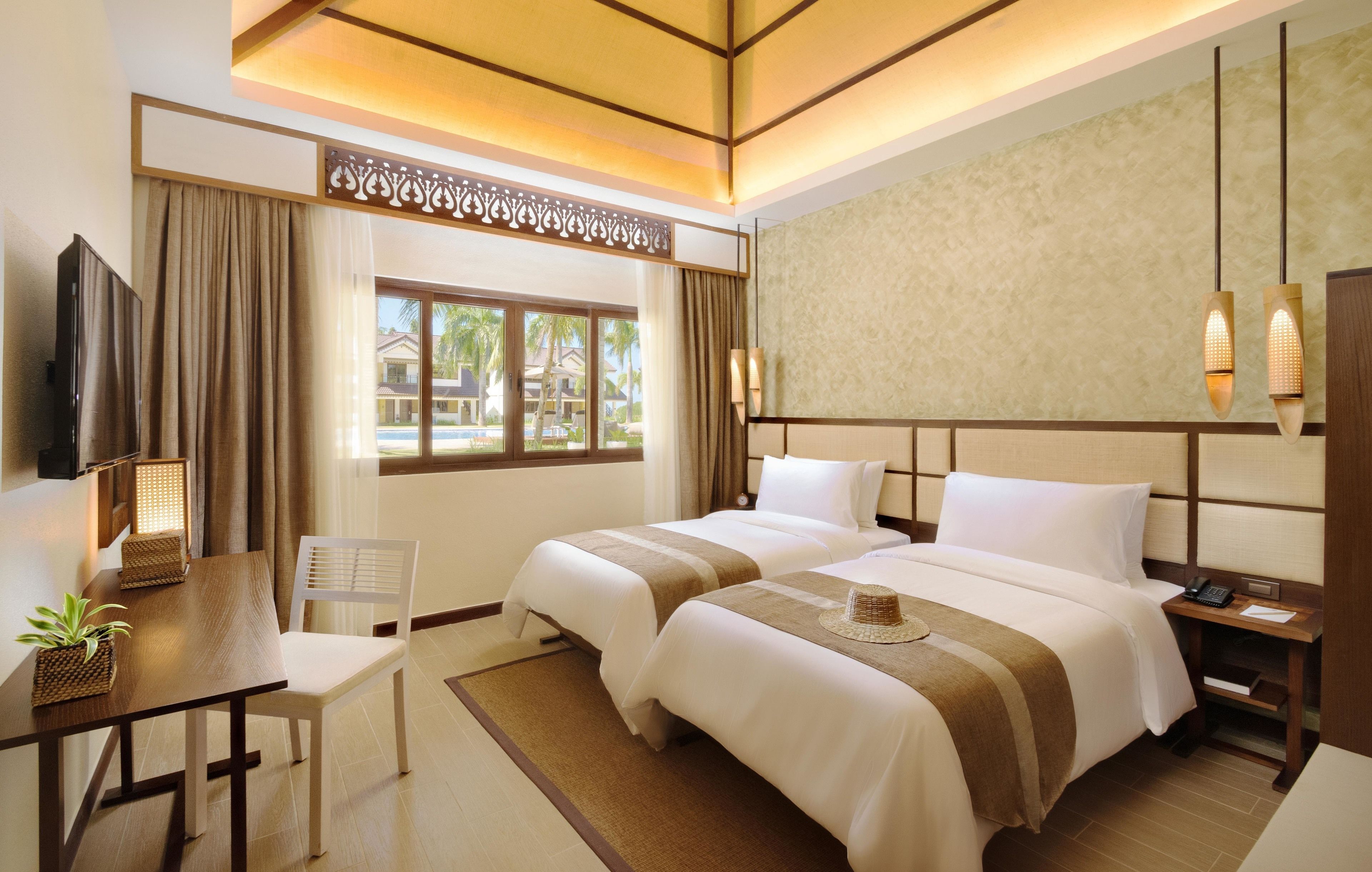 A hotel room with two beds, a desk with a chair, a TV, and a view of palm trees.