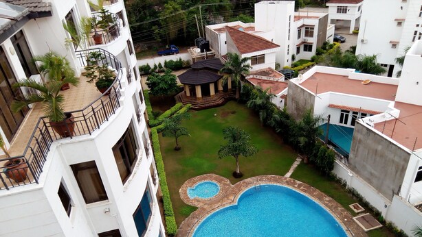Un área residencial con una amplia piscina rodeada por un borde de piedra, un pequeño jardín con un pabellón y un edificio blanco con balcones.