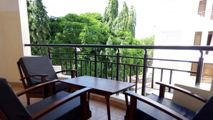 Un balcón con una mesa y cuatro sillas, suelo de baldosas y vistas a árboles verdes y un edificio al fondo.