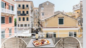 A balcony with wicker chairs, a table with a charcuterie board, and a bottle of wine. In the background, there are buildings with balconies and a hotel sign. - Nicol House and Suite in Corfu Town by Konnect  (Corfu)