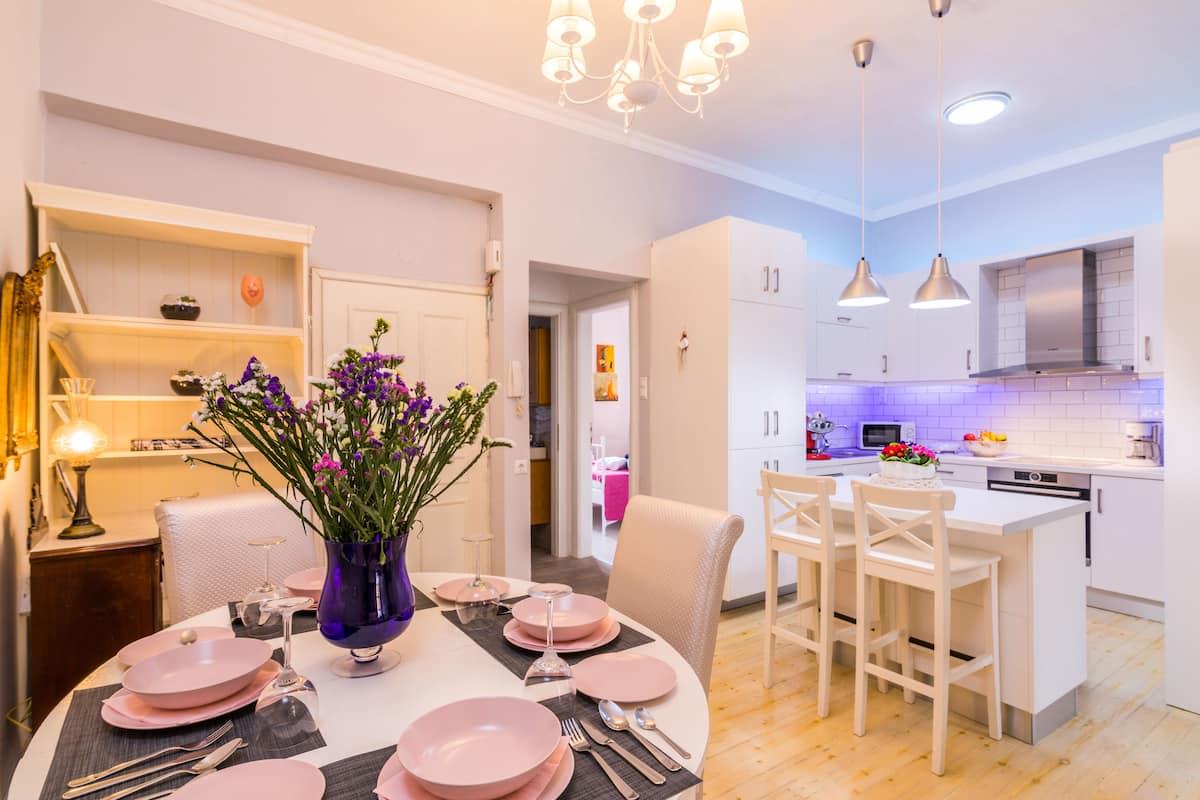 Una cocina moderna con armarios blancos, una isla central, iluminación colgante y una zona de comedor con vajilla rosa.