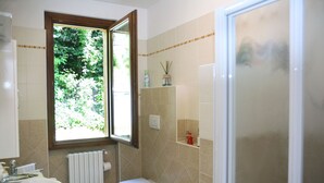 Badeværelse med toilet, bidet, vask og et vindue med udsigt over grøn vegetation.