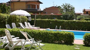 Et poolområde med liggestole og en parasol, omgivet af velplejede hække og en klar, blå pool.