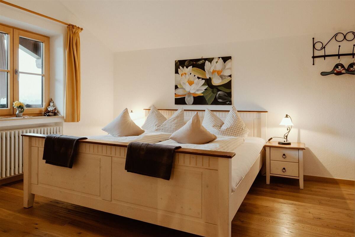 Quarto com cama de madeira, roupa de cama branca, mesa de cabeceira com abajur, janela com cortinas e prateleira na parede.