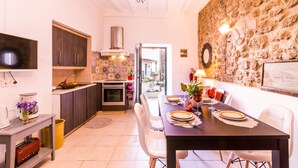 A modern kitchen with dark cabinets, a tiled backsplash, and a stone wall. There is a dining area with a table set for two, a chandelier, and a view of the outside. - Nina's Traditional House by Estia (Corfu)