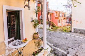 Vista da rua com uma mesa branca posta, com xícaras e uma fruteira, uma porta verde e um edifício com fachada vermelha ao fundo.