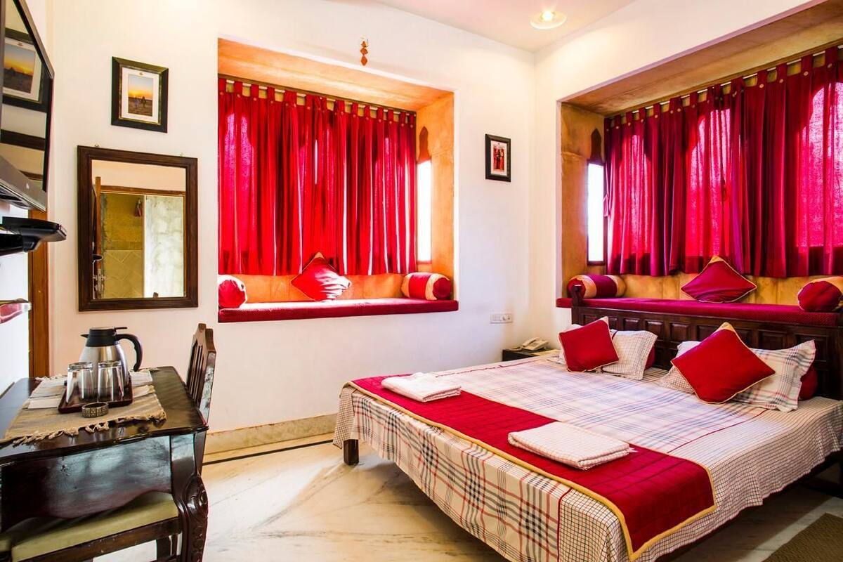 A room with a bed, a window with red curtains, a small table with a kettle, and a mirror on the wall.