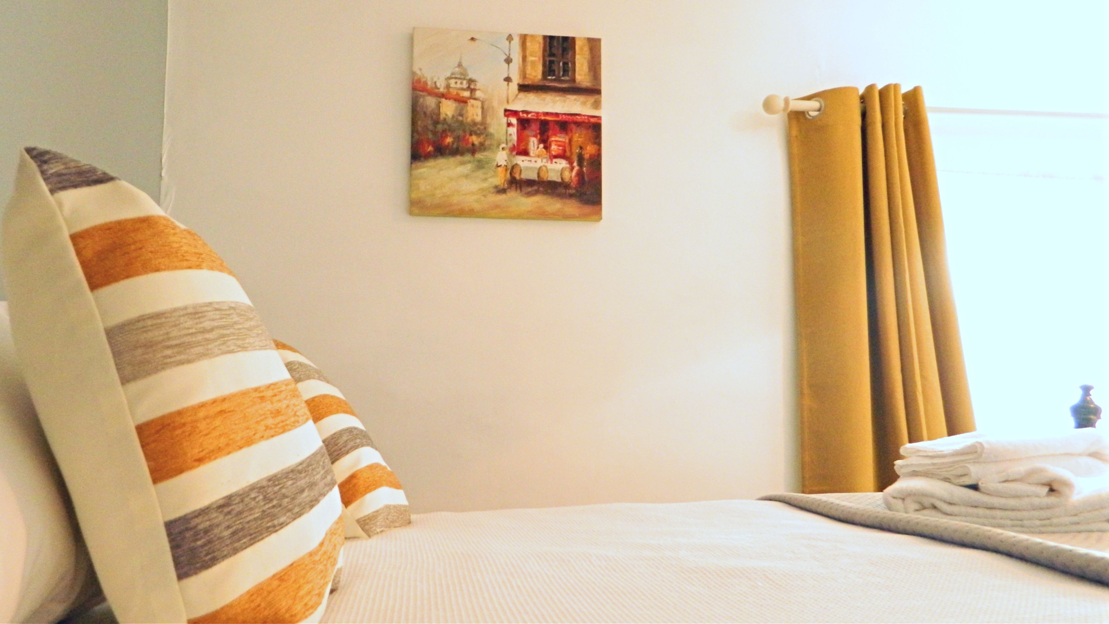 A hotel room with a bed, a pillow, a painting on the wall, and a window with curtains.