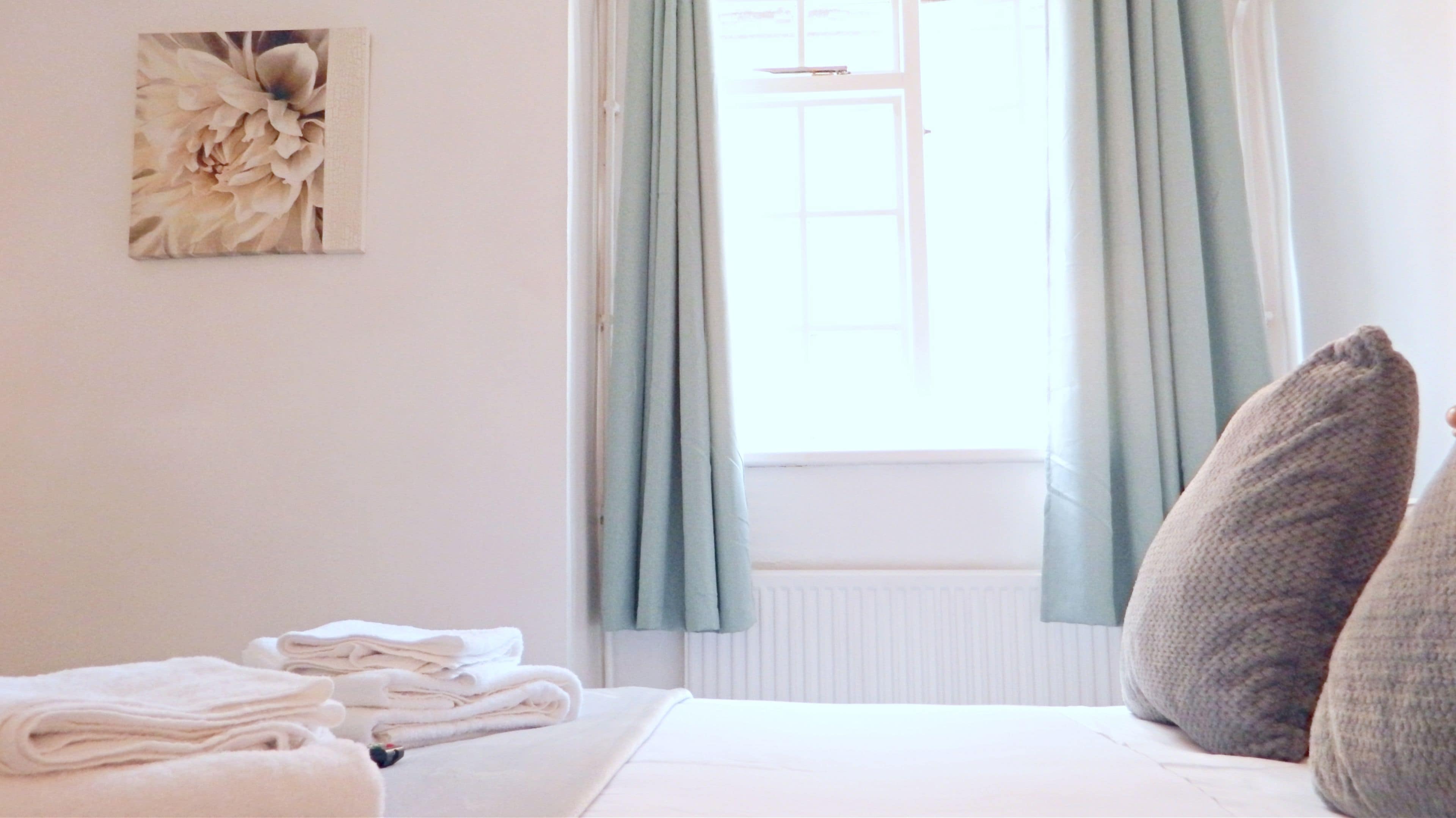 A neatly made bed with folded towels, a window with light blue curtains, and a floral artwork on the wall.