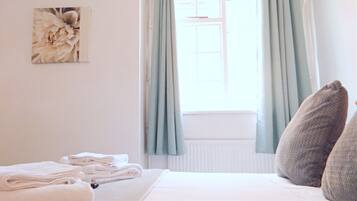 A neatly made bed with folded towels, a window with light blue curtains, and a floral artwork on the wall.