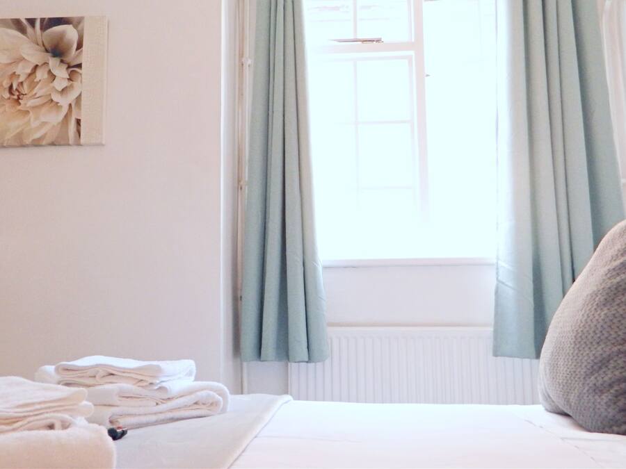 A neatly made bed with folded towels, a window with light blue curtains, and a floral artwork on the wall.