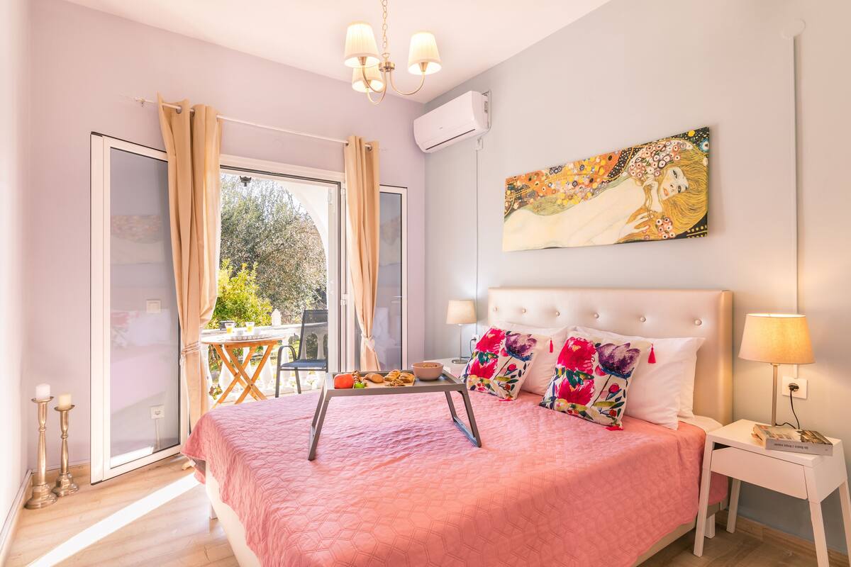Ein Schlafzimmer mit rosa Bettdecke, einem Nachttisch mit Lampe, einem Bild an der Wand und einem Fenster mit Vorhängen.